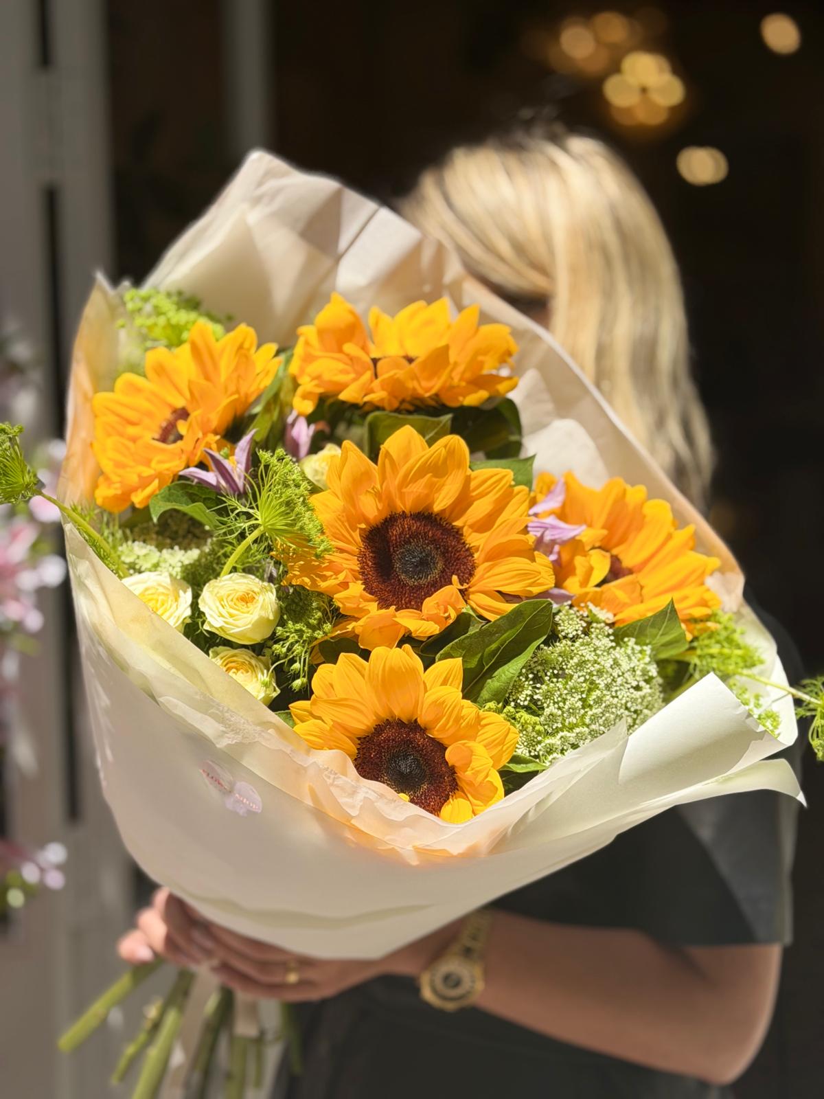 Ramo de girasoles amarillos, rosas spray blancas, clemátides lilas y follaje verde, envuelto en papel crema y sostenido por una mujer.