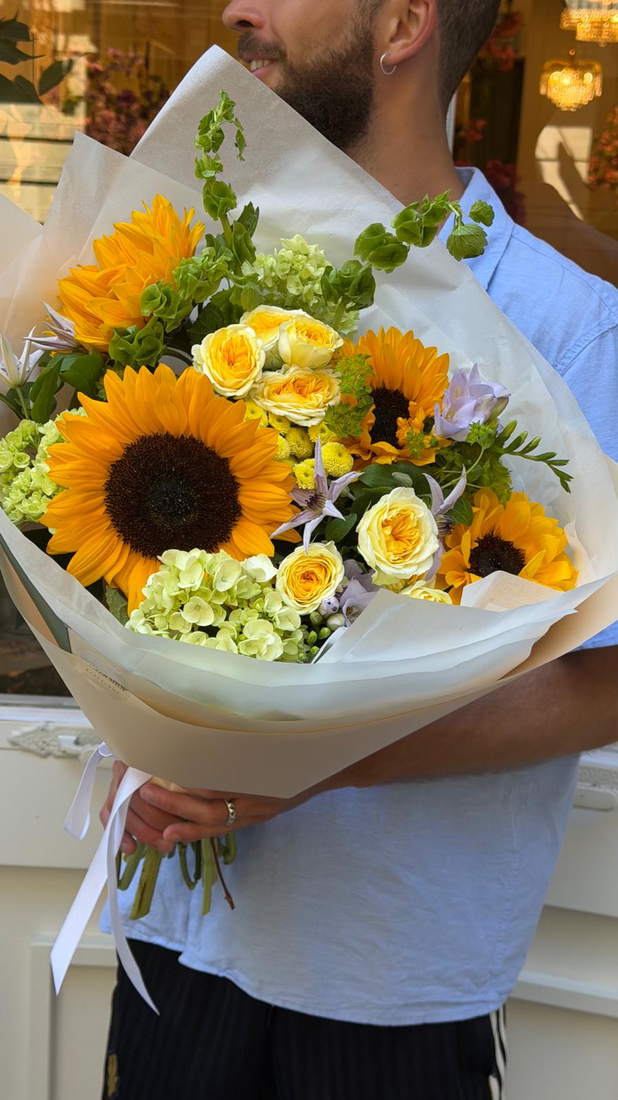 Un ramo alegre y luminoso con girasoles, rosas spray, ammi blanco y delicadas flores en tonos lila. Perfecto para cumpleaños, celebraciones o regalos especiales.