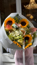 Ramo de girasoles amarillos, anturios rosa empolvado, hortensias blancas, rosas spray crema, rosas rosadas, solidago dorado y eucalipto, envuelto en papel blanco.