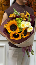 Person holding the Luz de Girasol bouquet, featuring large golden sunflowers, cream roses, fresh green blooms, purple accent flowers, and golden eucalyptus, wrapped in deep burgundy and beige paper with mauve ribbon.