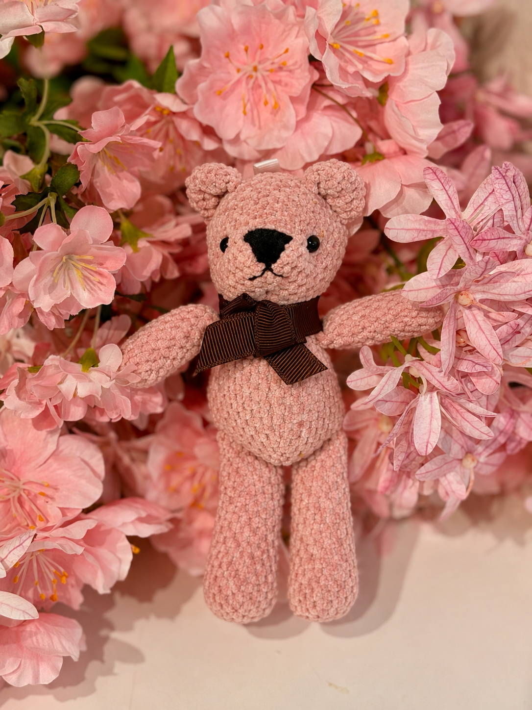 Oso de peluche pequeño rosa con lazo marrón entre flores rosas
