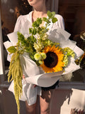 Ramo Petit Soleil con girasol amarillo, flores blancas, crisantemos verdes y amaranto colgante, envuelto en papel blanco.