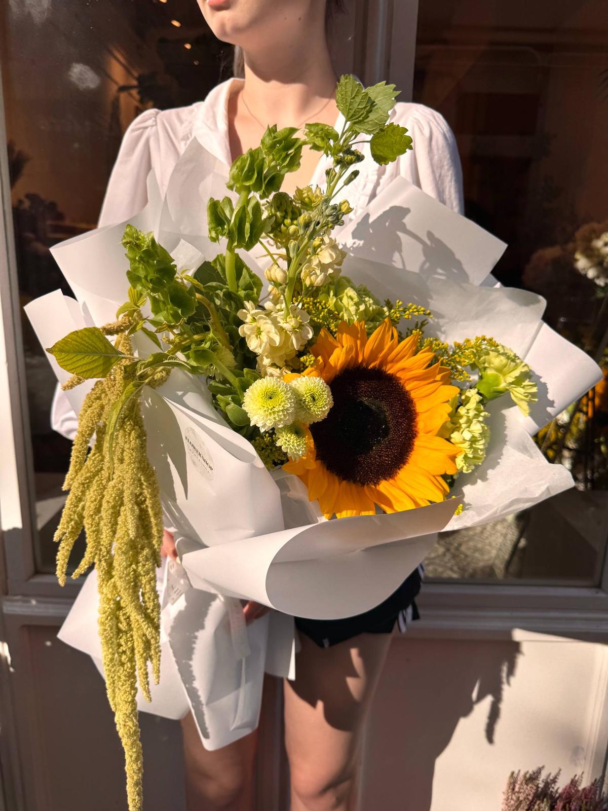 Ramo Petit Soleil con girasol amarillo, flores blancas, crisantemos verdes y amaranto colgante, envuelto en papel blanco.