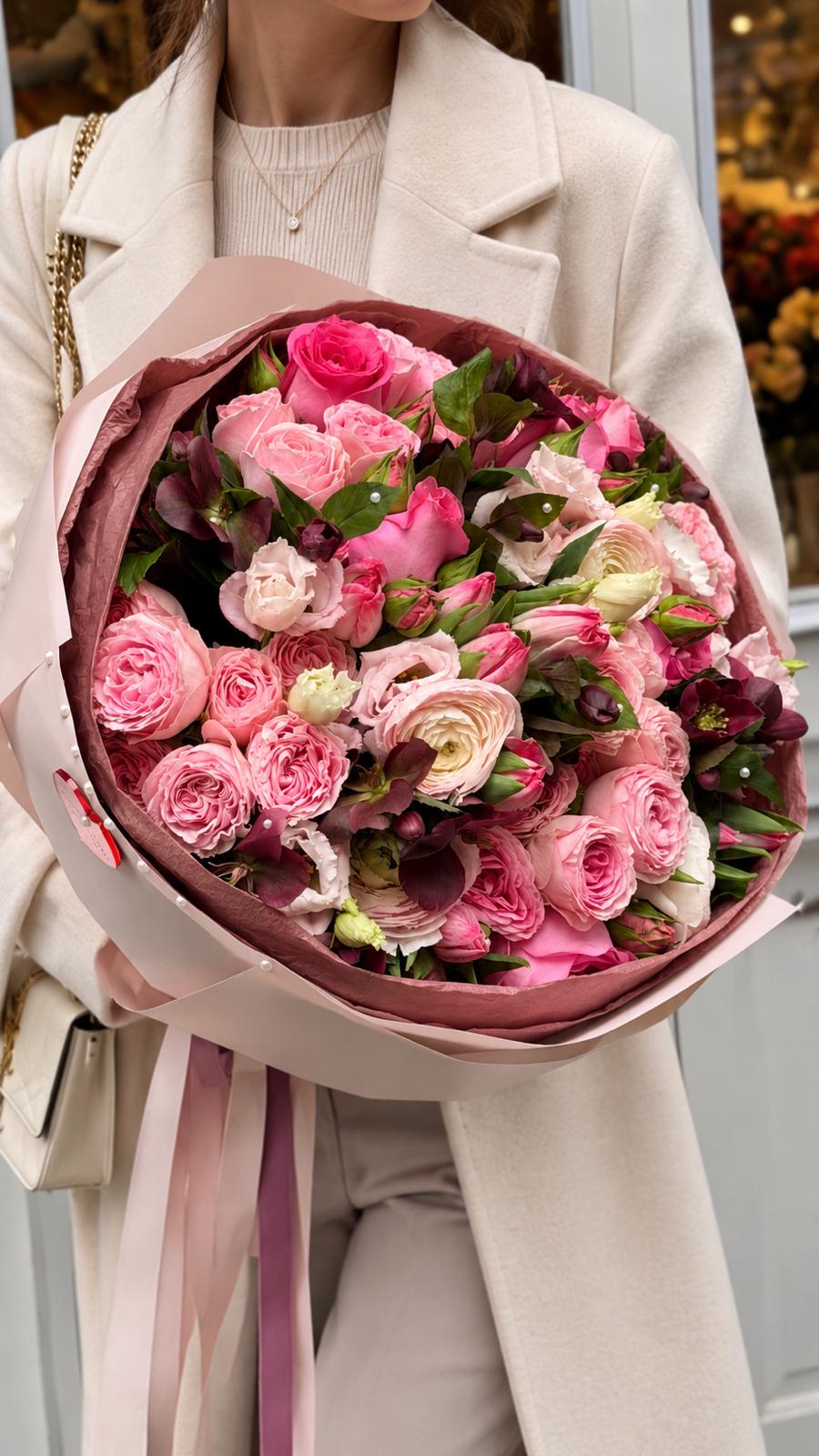 Ramo Rosa & Ranúnculo Frambuesa con rosas de jardín y ranúnculos en tonos rosa y frambuesa, lisianthus claros y eléboros borgoña, envuelto en papel rosa y sostenido por una mujer.
