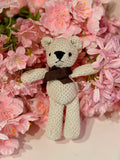 Oso de peluche pequeño blanco con lazo marrón entre flores rosas
